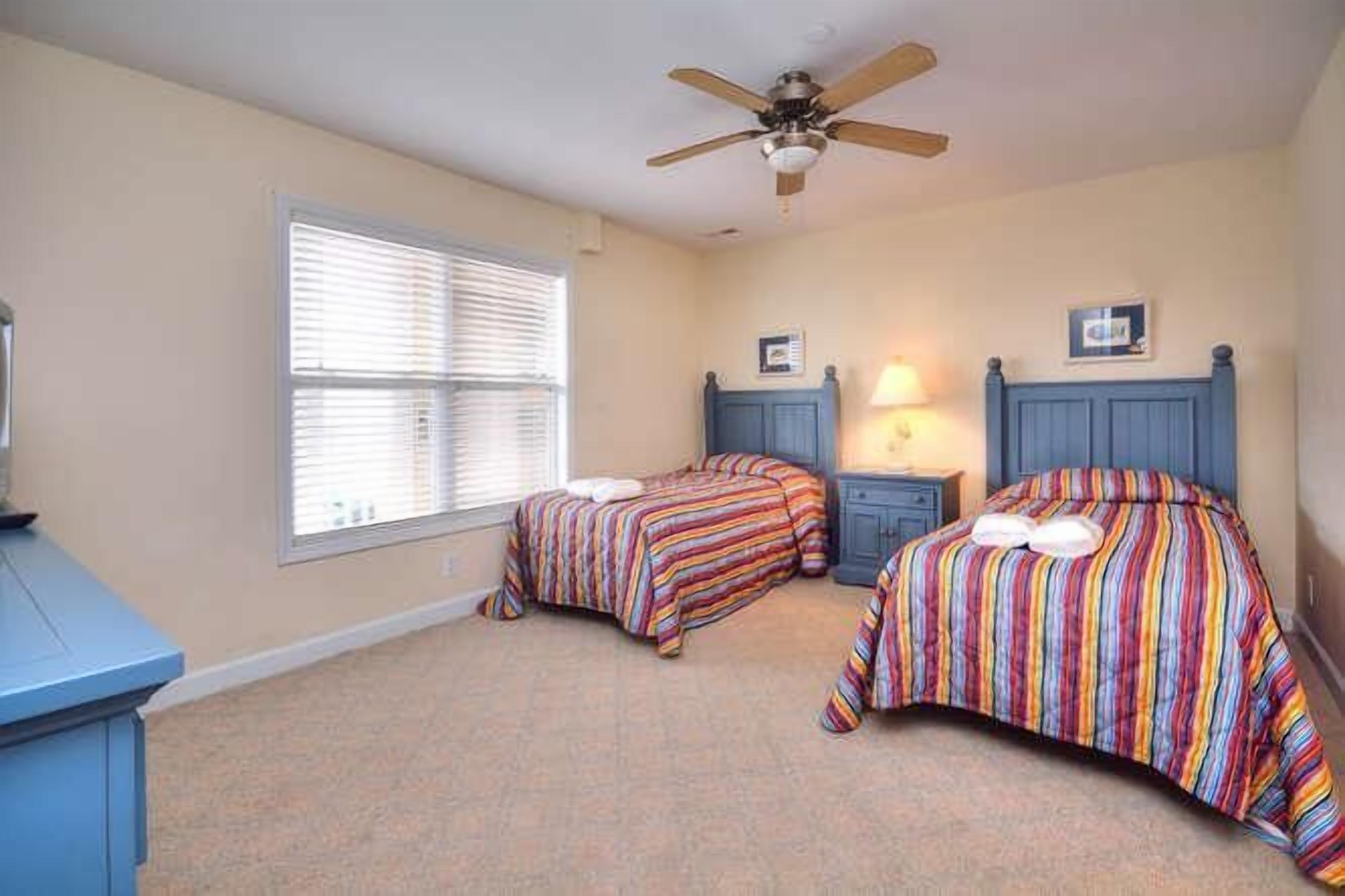A bedroom with two beds, a ceiling fan, a window with blinds, and a desk.