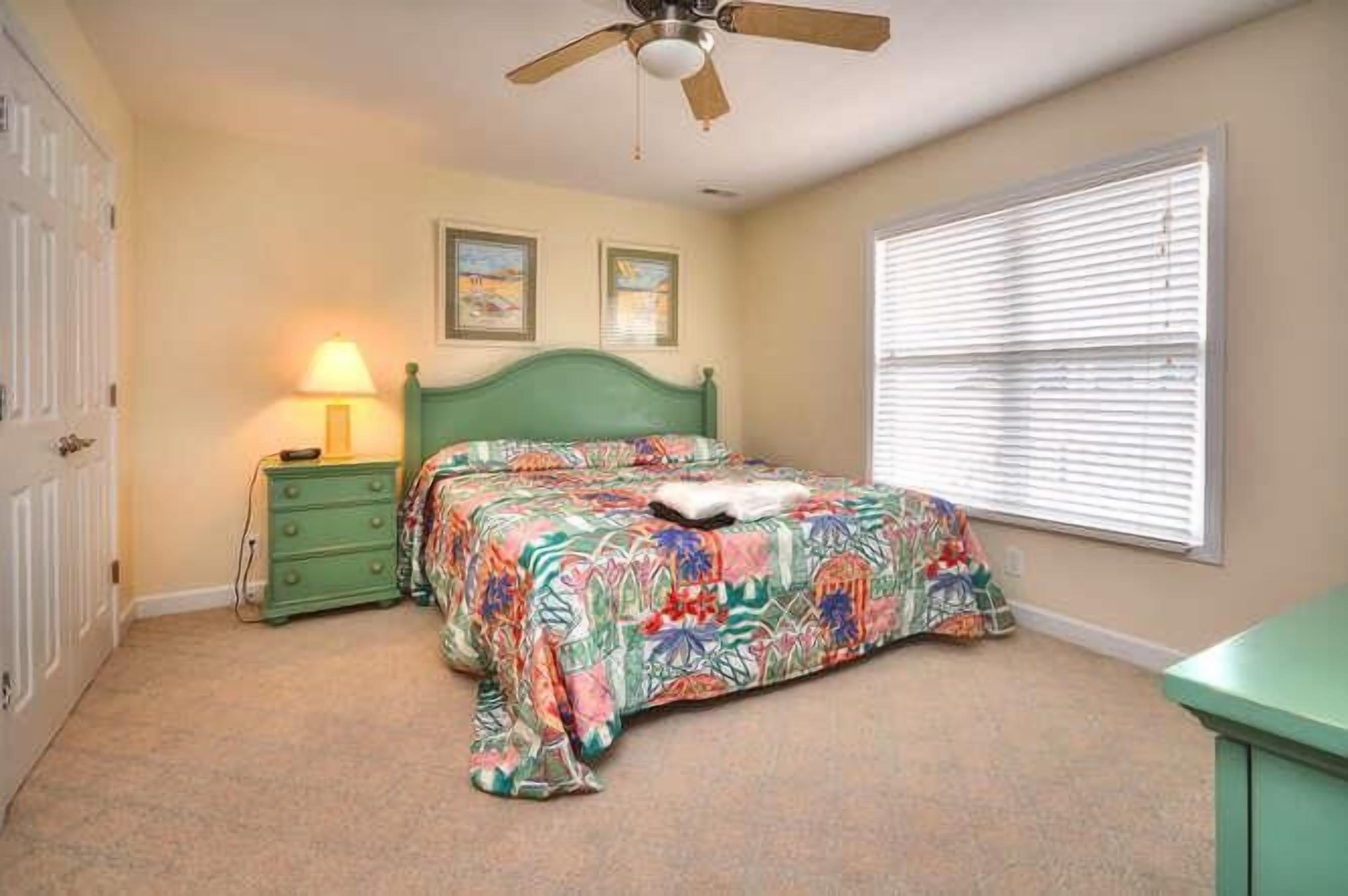 A bedroom with a bed, a nightstand, a lamp, a ceiling fan, and a window with blinds.