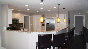 A kitchen with white cabinets, a stainless steel refrigerator, and a granite countertop. There are two bar stools and pendant lights above the counter. - Have it All!!! True Water Front, Amazing Views (Golden)