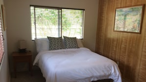 A neatly made bed with white bedding, two patterned pillows, a bedside table with a lamp, and a window with blinds.