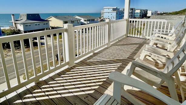 Une terrasse en bois avec des balustrades et des chaises blanches, donnant sur une plage où l’on aperçoit des maisons au loin.
