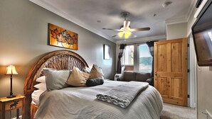 A bedroom with a large bed, a ceiling fan, a wooden headboard, a painting of leaves, and a wooden door.