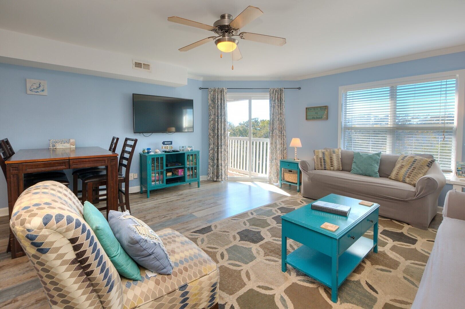 A living room with a sofa, coffee table, and patterned armchair. There's a dining area with a table and chairs, a TV, and a ceiling fan.