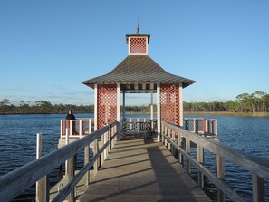 Eine hölzerne Mole führt zu einem Pavillon mit Gitterverkleidung, mit Blick auf ruhiges Wasser.