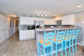 Una cocina con armarios blancos, una isla central con sillas azules y un refrigerador de acero inoxidable.