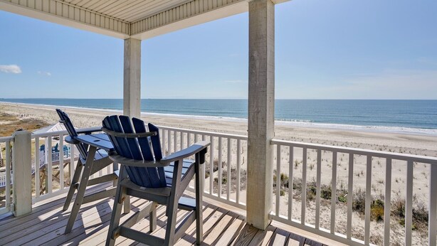A covered deck with a view of the beach, a table, and two chairs. - Discover oceanfront bliss at 7th Heaven in Oak Island, NC—spacious 7BR, sleeps 24! (Oak Island)