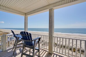 A covered deck with a view of the beach, a table, and two chairs. - Discover oceanfront bliss at 7th Heaven in Oak Island, NC—spacious 7BR, sleeps 24! (Oak Island)