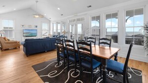 Un salon spacieux avec un grand canapé, une table à manger et des chaises, un ventilateur de plafond et de grandes fenêtres offrant une vue sur l’extérieur.