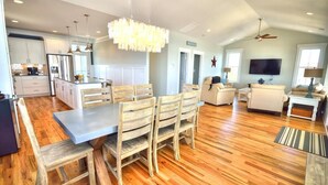 Un'ampia zona giorno con un tavolo da pranzo, sedie in legno, un lampadario, una ventola a soffitto, una televisione e una cucina sullo sfondo.