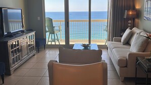 A living room with a television, a sofa, a chair, and a table. There is a view of the ocean through large sliding doors.