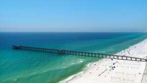 A beach with a long pier extending into the ocean, a sandy beach area with umbrellas, and a clear blue sky.