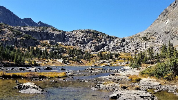A mountainous landscape with a clear stream, rocky terrain, and dense forest.