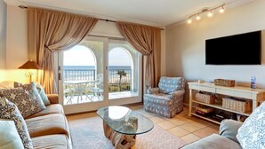 A living room with a sofa, armchair, and glass coffee table. There is a TV mounted on the wall and a balcony with curtains. - Beautiful 2br/2ba Oceanfront, Sea Dunes  (Amelia Island)