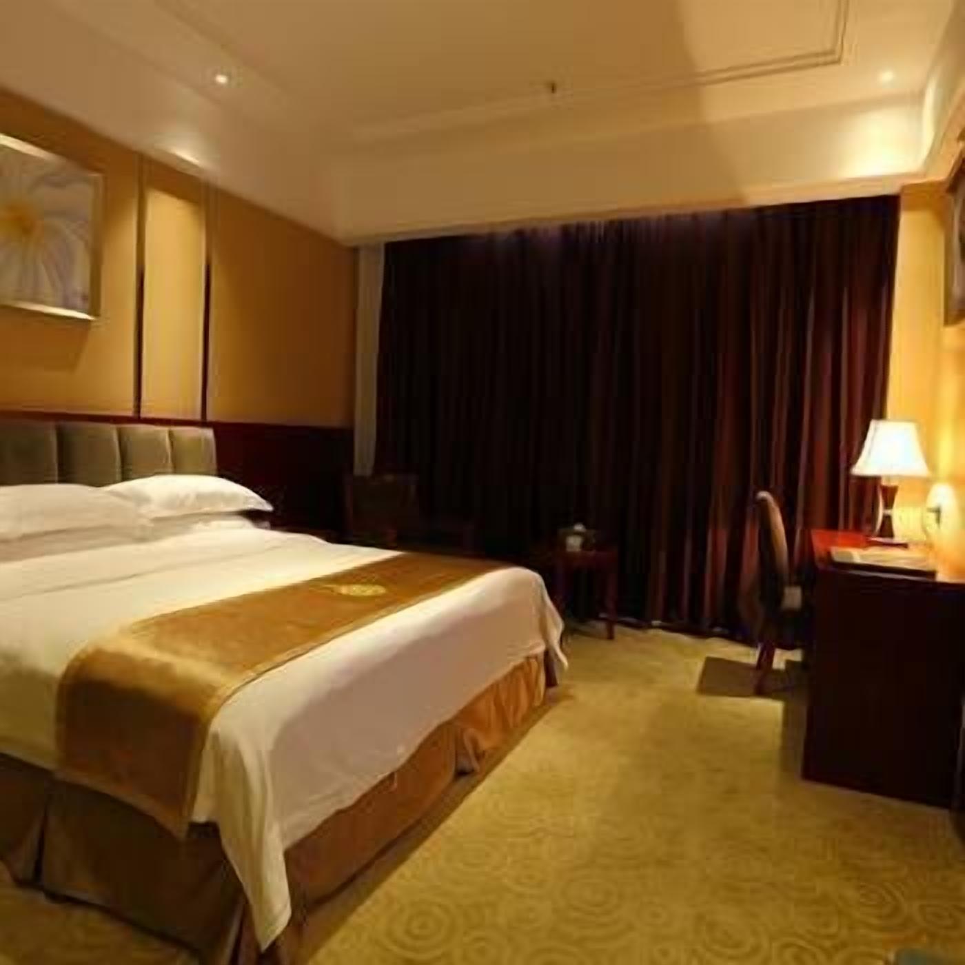 A hotel room with a large bed, a desk with a lamp, and a window with curtains.