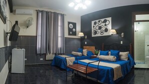 Habitación con dos camas, un escritorio, un refrigerador, un ventilador de pared y una puerta que da al baño.