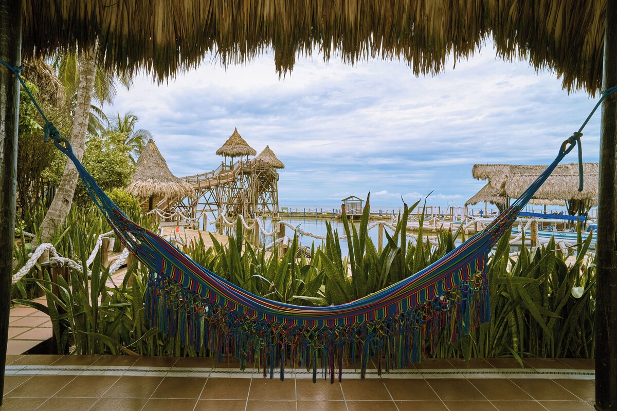A hammock in a tropical setting with thatched-roof structures and a view of the ocean.