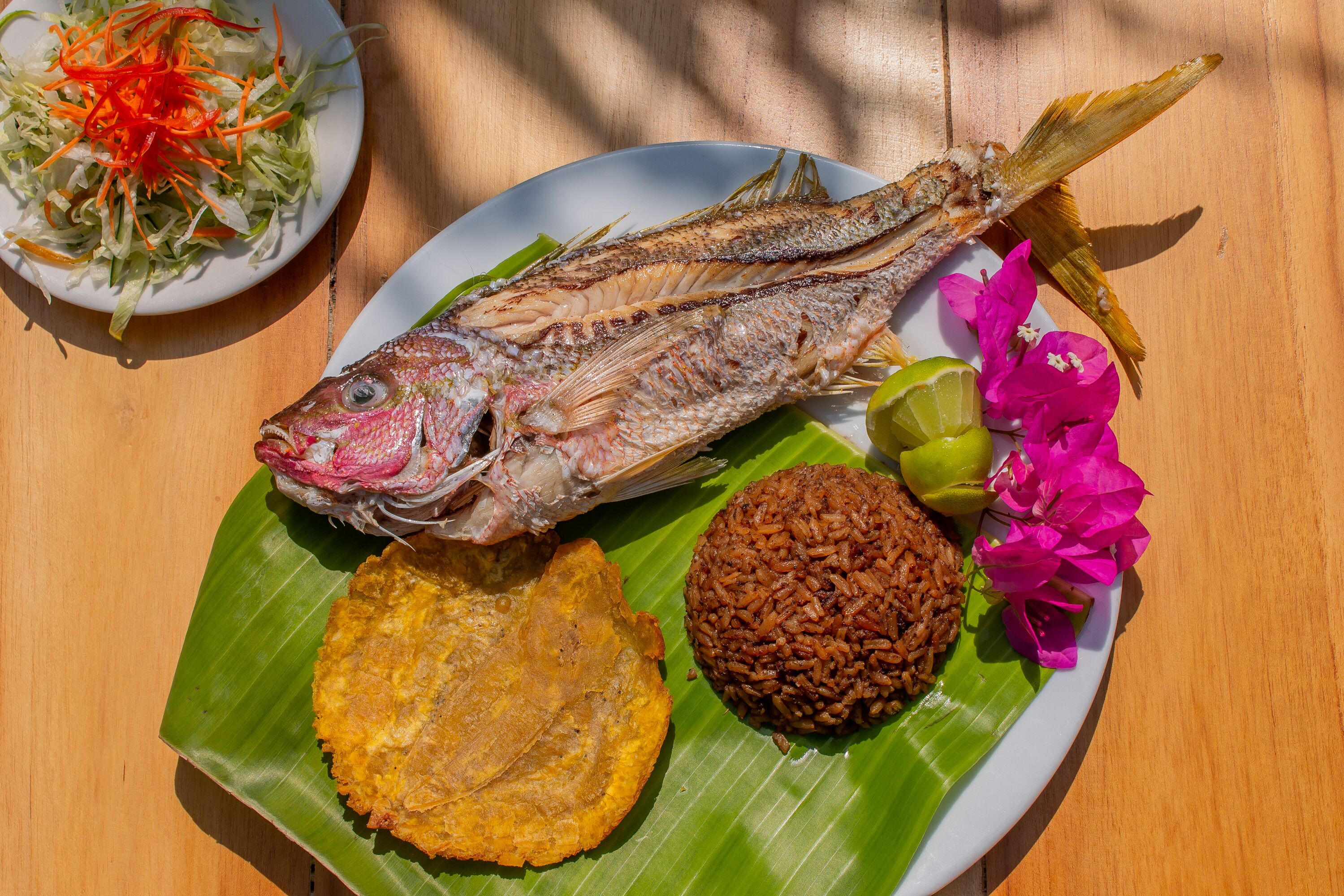 Ein Teller mit Essen, darunter ein ganzer Fisch, Reis und gebratene Kochbananen auf einem Bananenblatt.