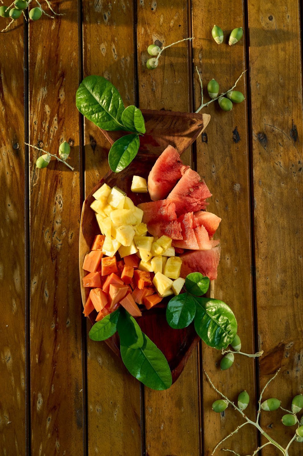 A wooden table with a wooden bowl containing diced fruits and green leaves.