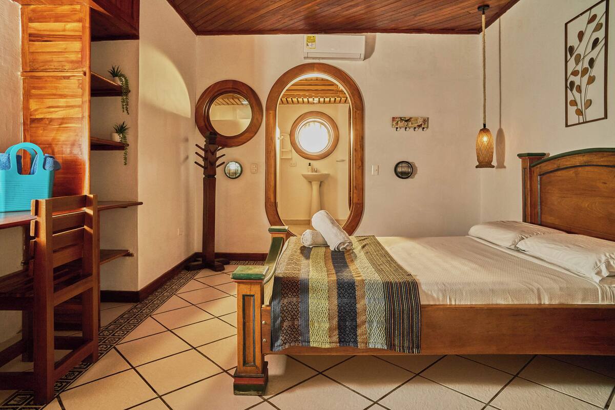 A wooden bed with a striped blanket, a wooden desk with a blue bag, a mirror, and a sink in a room with tiled flooring.