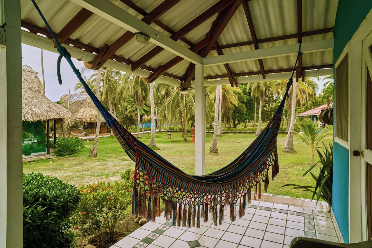 A hammock under a covered area with a tiled floor, palm trees, and a thatched-roof structure in the background.