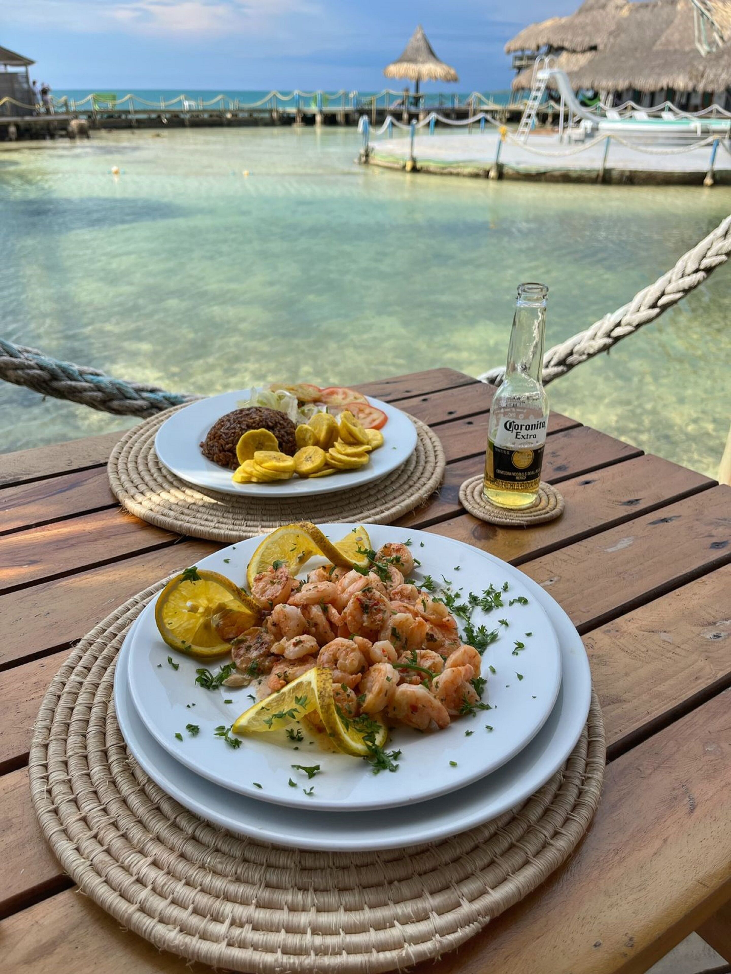 Deux assiettes de nourriture sur une table en bois, avec en arrière-plan une vue sur une mer bleue et dégagée et une construction au toit de chaume.