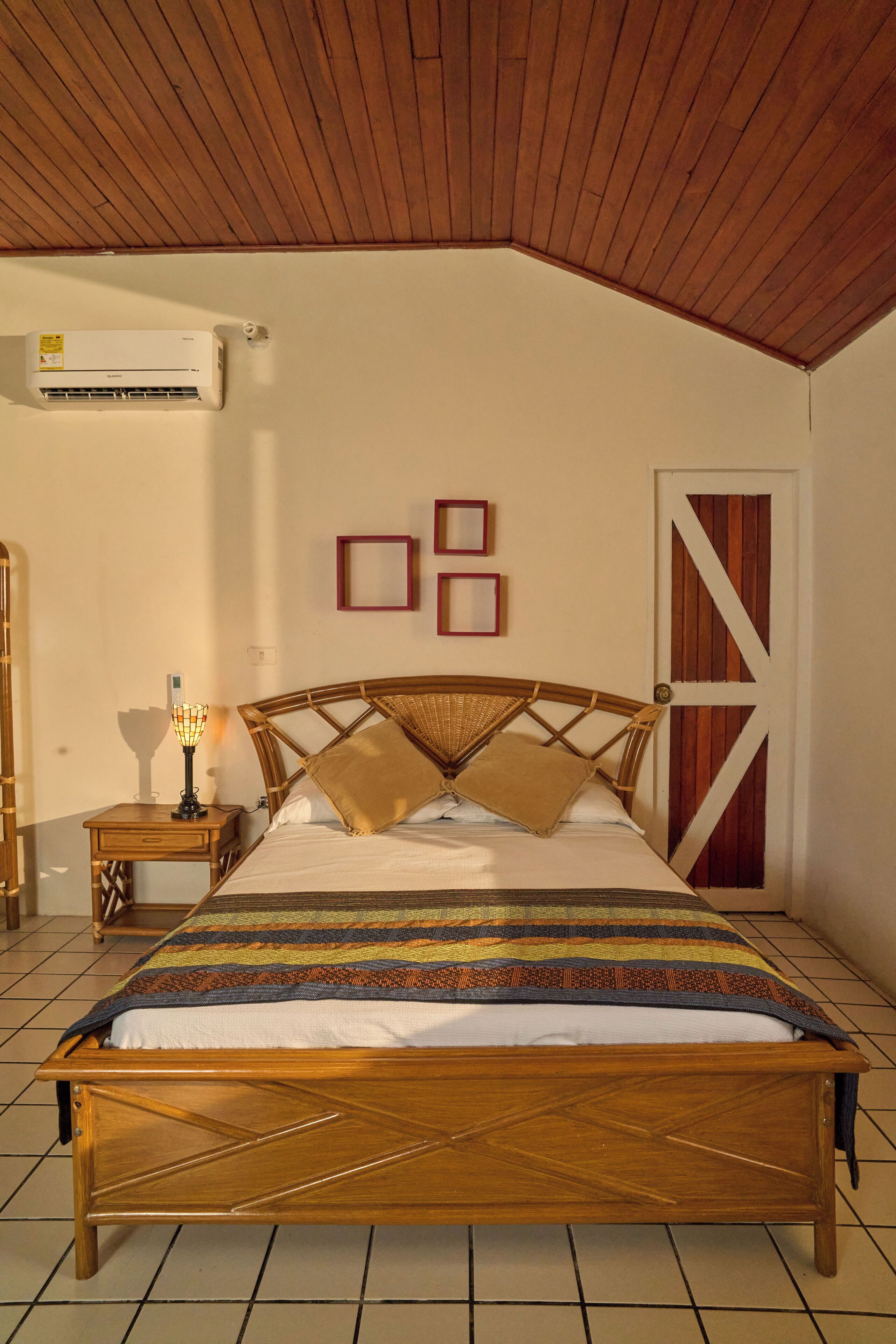 A bedroom with a wooden ceiling, a bed with a patterned bedspread, a nightstand with a lamp, and a wall-mounted air conditioner.