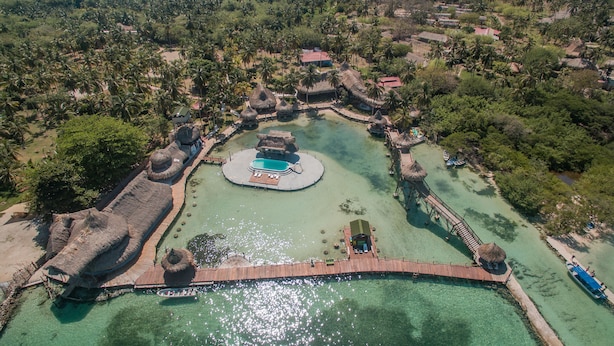 An aerial view of a resort with a central pool, thatched-roof structures, and a wooden bridge.
