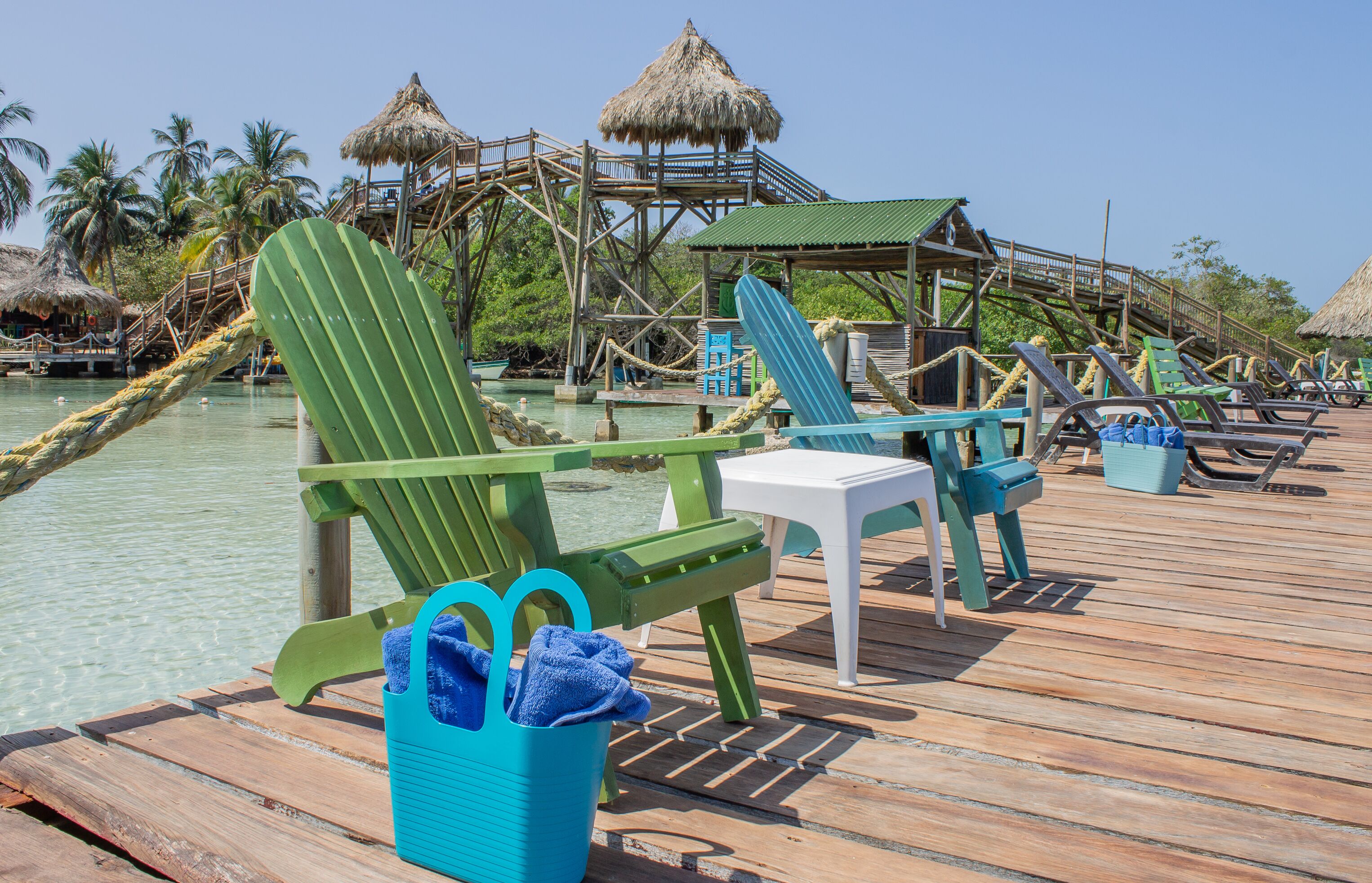 A wooden deck with green and white lounge chairs, a small table, and a blue bucket with a towel.