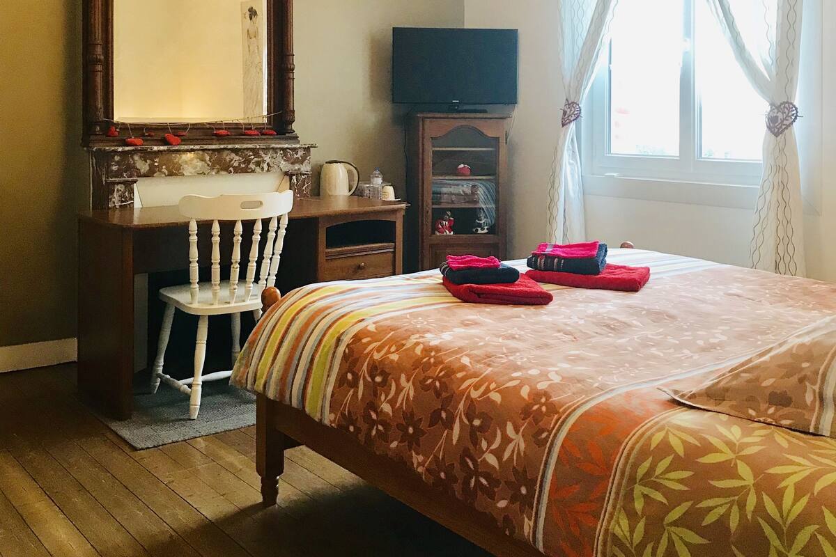 A hotel room with a wooden desk, a chair, a television, a bed with floral bedding, and a window with curtains.