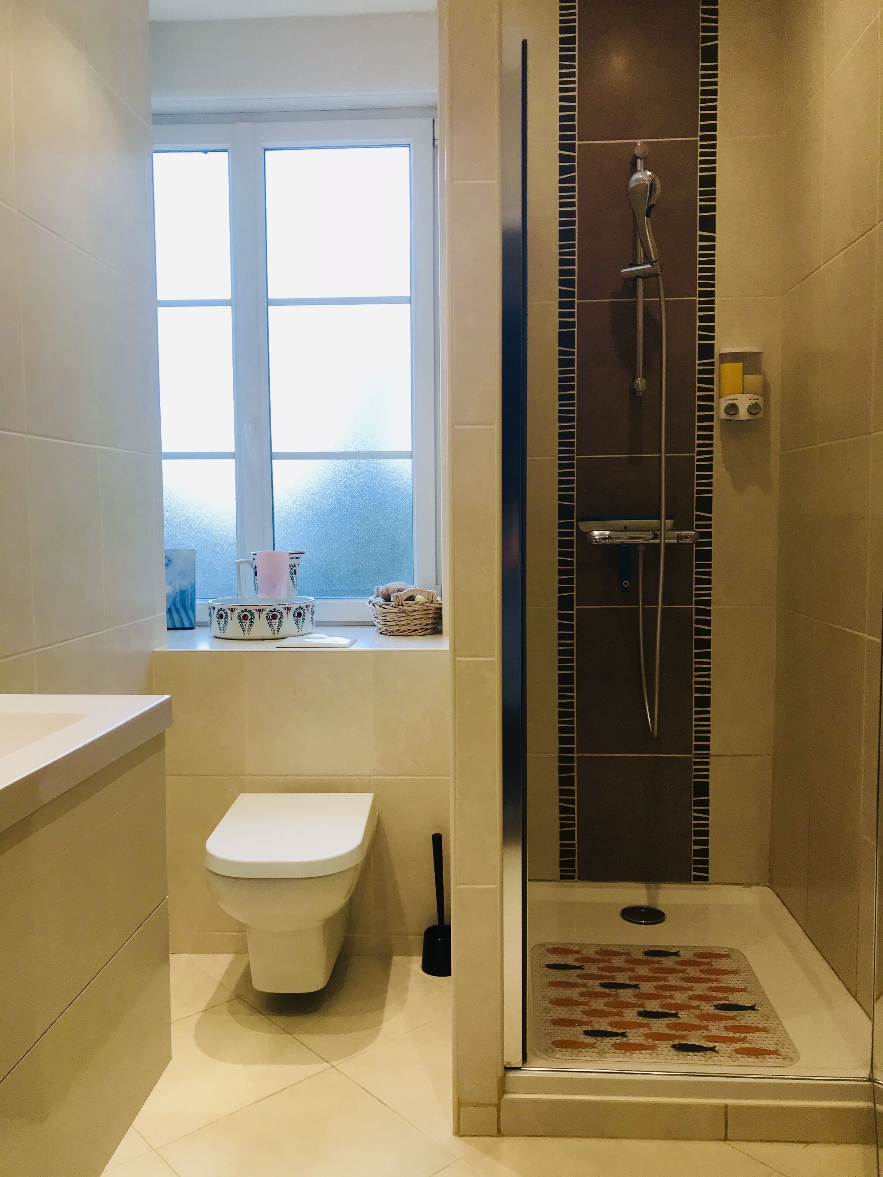 A bathroom with a toilet, a shower area with a glass door, a window, and a sink with a decorative bowl.