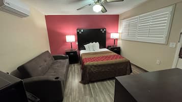 A hotel room with a bed, two bedside tables with lamps, a ceiling fan, a window with blinds, a sofa, and a desk.