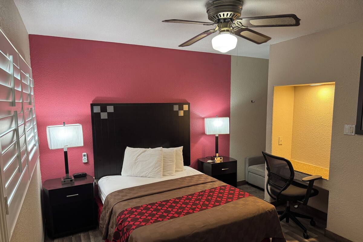A hotel room with a bed, two bedside tables with lamps, a desk with a chair, a ceiling fan, and a window with a view.