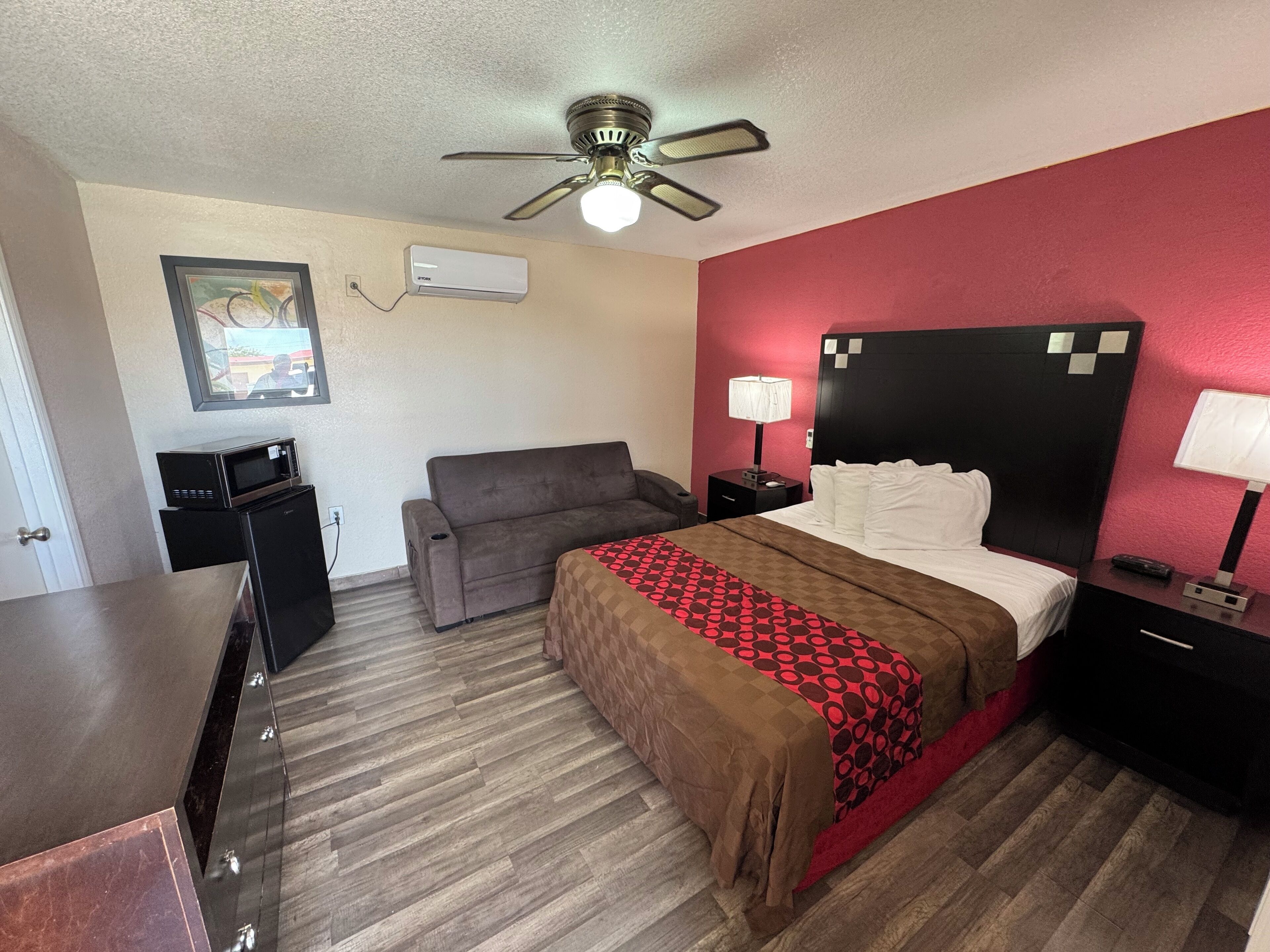 Una habitación de hotel con cama, escritorio, silla, ventilador de techo, mesita de noche y lámpara.