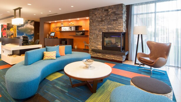 A modern living room with a curved blue sofa, a round coffee table, a stone fireplace, and a large window with sheer curtains. - Fairfield Inn & Suites by Marriott Buffalo Amherst/University (Buffalo)