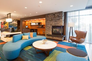 A modern living room with a curved blue sofa, a round coffee table, a stone fireplace, and a large window with sheer curtains. - Fairfield Inn & Suites by Marriott Buffalo Amherst/University (Buffalo)
