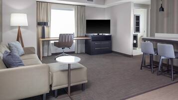 A modern hotel room with a sofa, a small round table, a TV, and a bar area with two chairs.