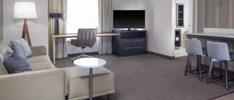 A modern hotel room with a sofa, a small round table, a TV, and a bar area with two chairs.