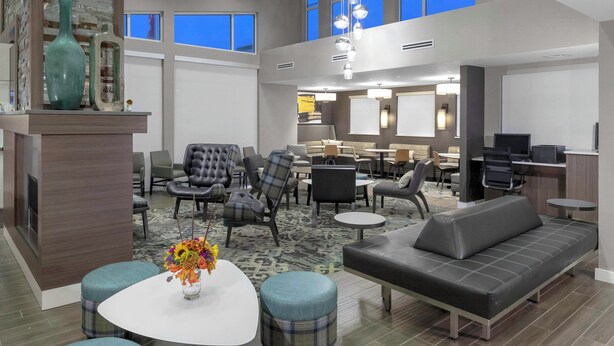 Un amplio vestíbulo de hotel con diseño moderno, que cuenta con una zona de estar con sofás y sillones, una mesa de centro con un jarrón de flores, y un gran ventanal con vistas al cielo.