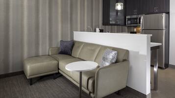 A modern kitchenette with a stainless steel refrigerator, microwave, and sink. A beige sectional sofa with a round coffee table is positioned in front of a large window with vertical blinds.