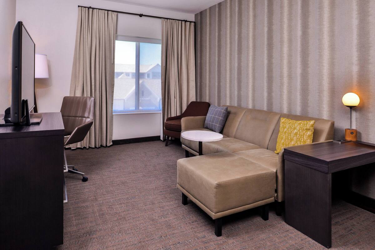 Habitación de hotel con sofá, una mesita redonda, escritorio con computadora, silla y lámpara.