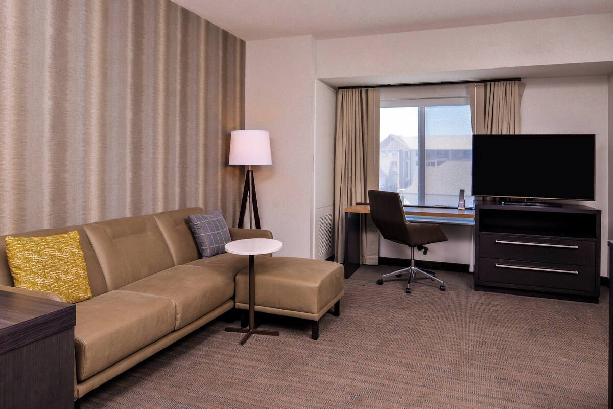 Habitación de hotel con sofá, una pequeña mesa redonda, escritorio con silla, televisión y un ventanal grande con cortinas.