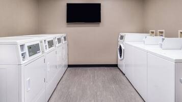 A laundry room with multiple washing machines and dryers, a flat-screen TV mounted on the wall, and a light-colored floor.