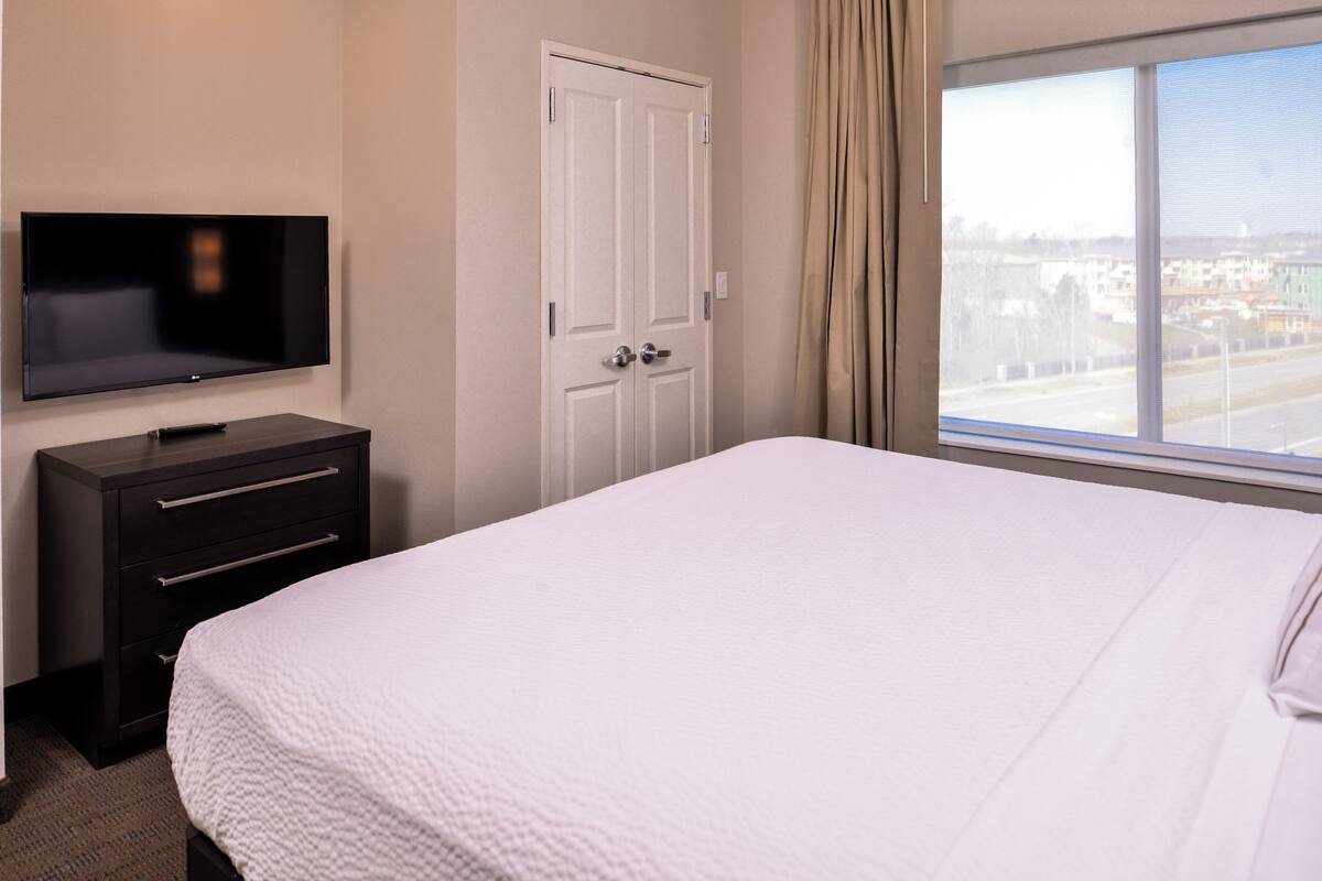 Habitación de hotel con cama, televisor en una cómoda, una puerta blanca y ventana con vistas a edificios.