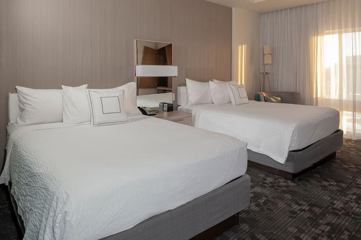 A hotel room with two beds, a bedside table with a lamp, a mirror, and a large window with curtains.