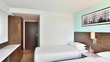 Habitación de hotel con dos camas, un armario de madera, un cuadro enmarcado de un paisaje urbano y una mesita de noche con un jarrón de flores.