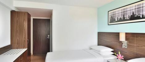 Habitación de hotel con dos camas, un armario de madera, un cuadro enmarcado de un paisaje urbano y una mesita de noche con un jarrón de flores.