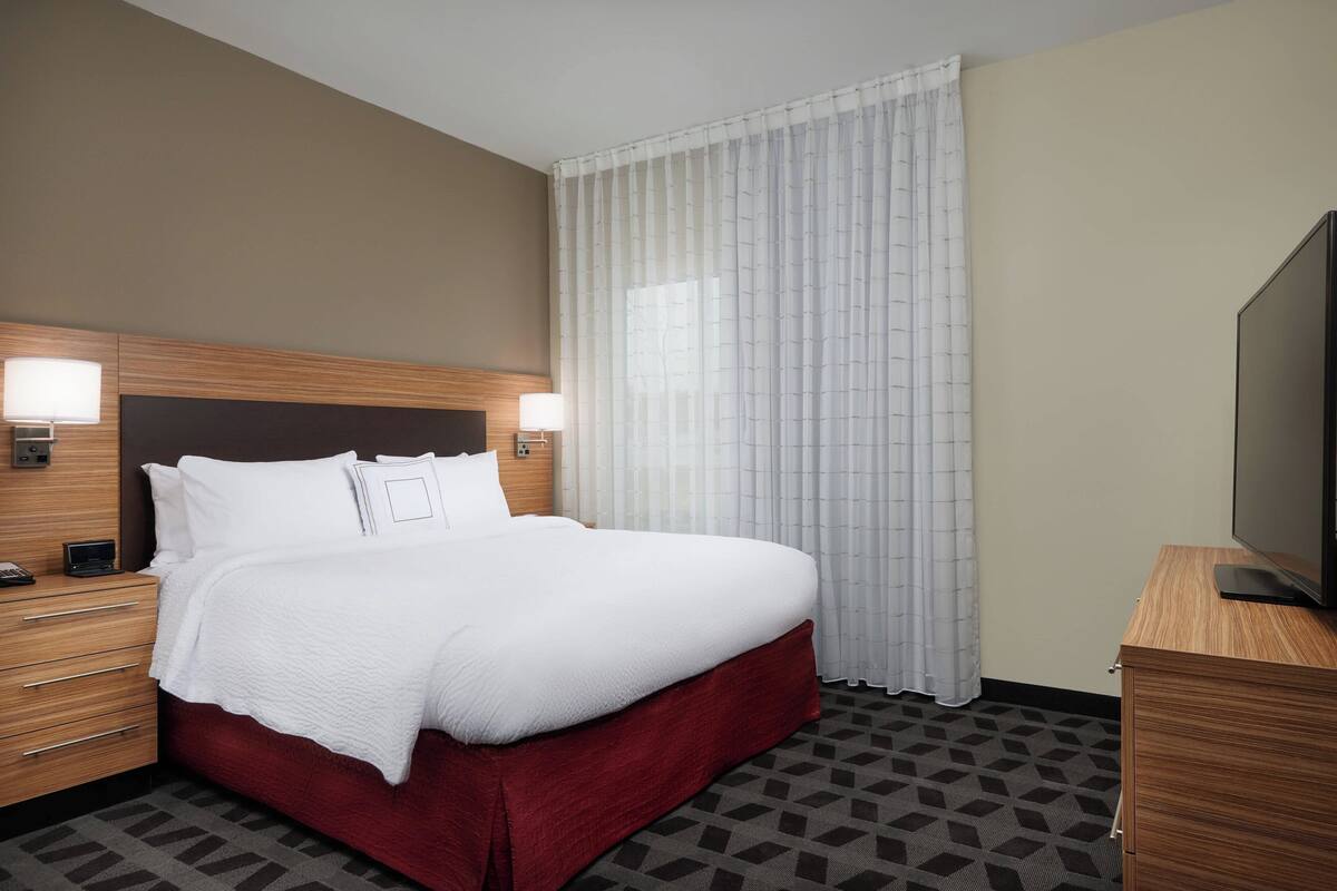 A hotel room with a large bed, two bedside tables with lamps, a flat-screen TV, and a window with sheer curtains.