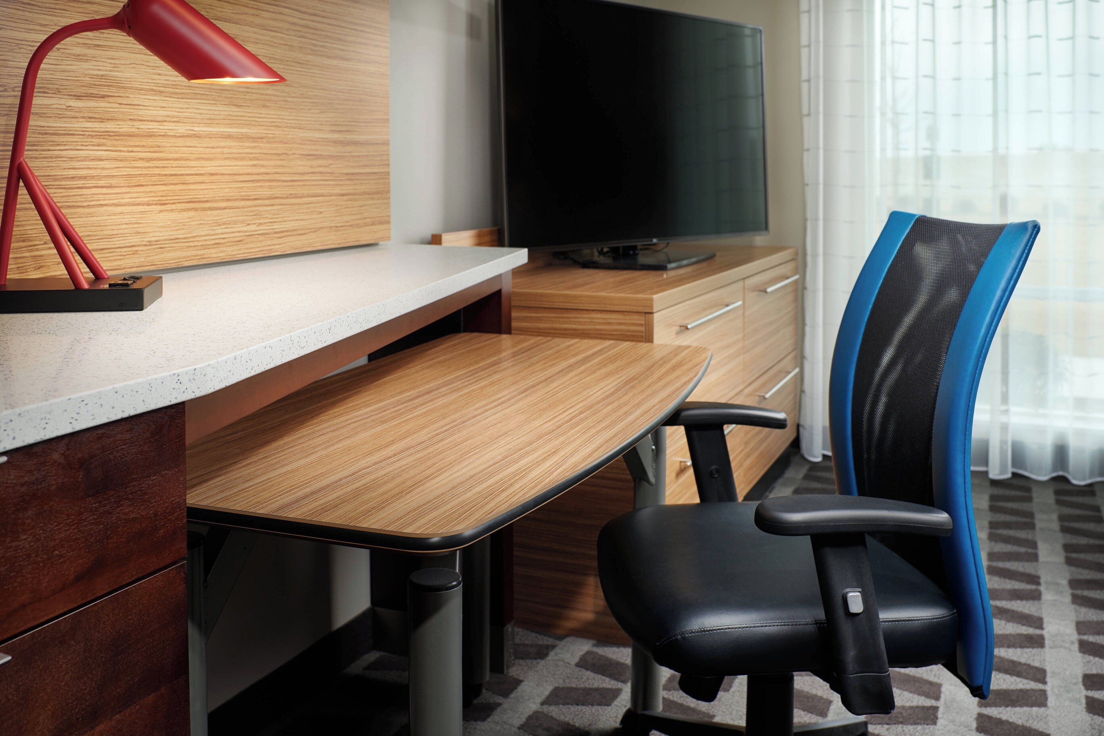 Une chambre d’hôtel moderne avec un bureau en bois, un fauteuil de bureau noir, une lampe de bureau rouge, un téléviseur à écran plat et une grande fenêtre ornée de rideaux légers.