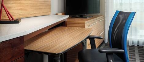 Une chambre d’hôtel moderne avec un bureau en bois, un fauteuil de bureau noir, une lampe de bureau rouge, un téléviseur à écran plat et une grande fenêtre ornée de rideaux légers.