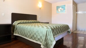 Uma cama bem arrumada com colcha xadrez num quarto com piso de madeira, luminária na parede e uma porta.